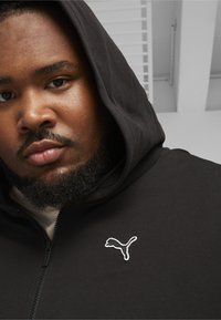 Sort zip-up hoodie med en blød tekstur og et hvidt Puma-logo på brystet. Hættens er trukket op og rammer ansigtet.