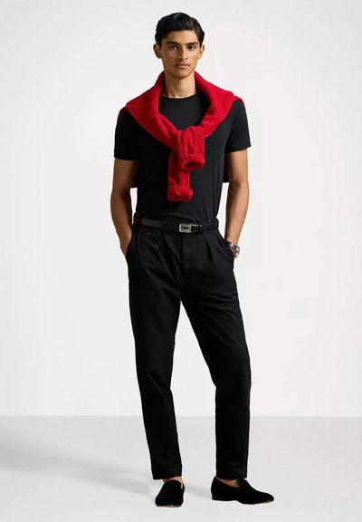 Pantaloni neri, t-shirt nera, maglione rosso legato attorno alle spalle e scarpe nere. Il modello è in piedi con le mani in tasca su uno sfondo neutro.