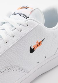 Sneaker alb din piele cu suprafață texturată, având un logo brodat negru și portocaliu și șireturi albe, cu cusături evidențiate.