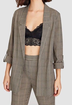 Femme portant un blazer marron à carreaux et un pantalon assorti avec un bralette en dentelle noire en dessous, montrée du cou aux hanches.