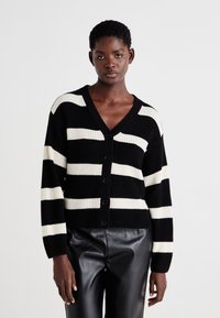 Zwart-witte gestreepte cardigan met een V-hals, knoopsluiting en ribbeltextuur. Draagt over zwarte leren broek, met lange mouwen.