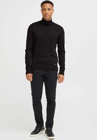 Maglione a collo alto nero, tessuto a maglia, design aderente, indossato con jeans neri e sneakers alte, modello in piedi su uno sfondo neutro.