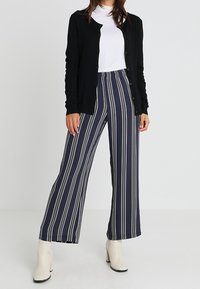 Cardigan nero, t-shirt bianca, pantaloni a vita alta a righe blu e bianche, stivali alla caviglia crema. Maniche lunghe, chiusura a bottoni sul cardigan.