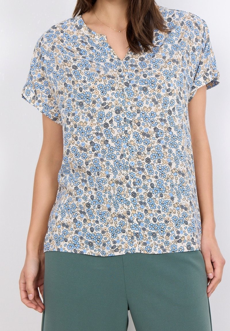 Femme portant un chemisier à manches courtes avec un motif floral bleu et beige et un pantalon vert, debout devant un fond uni.