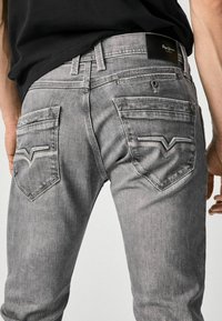 Jeans de mezclilla gris con un corte ajustado, con una etiqueta de cuero negro en la cintura y costuras decorativas en los bolsillos traseros.