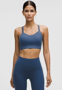 Mujer con un sujetador deportivo azul a juego con doble tirante y leggings de cintura alta, de pie frente a un fondo claro y liso.