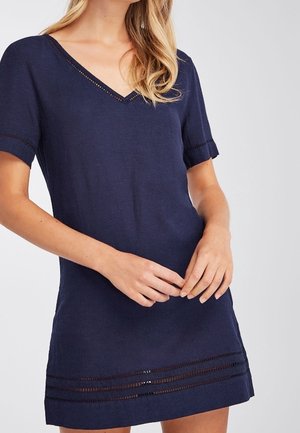 Mujer vestida con un vestido de manga corta azul marino con escote en V y detalles de cortes horizontales cerca del dobladillo, de pie con las manos suavemente entrelazadas.