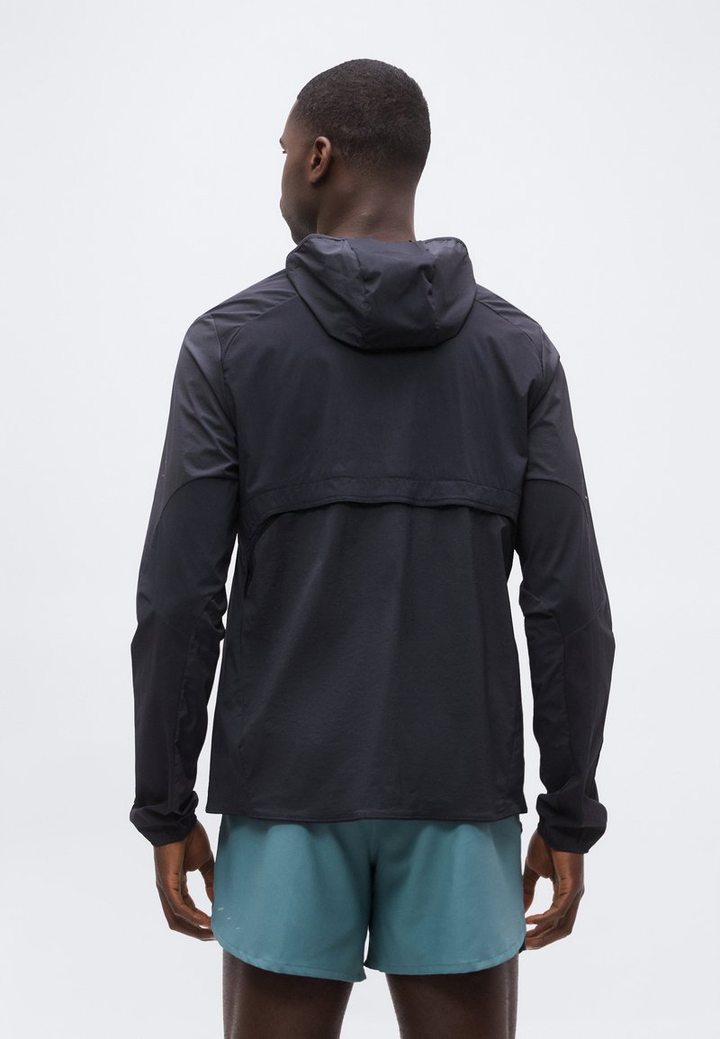 Hombre con una chaqueta negra con capucha y pantalones cortos deportivos de color azul verdoso, de pie de espaldas contra un fondo claro y liso.