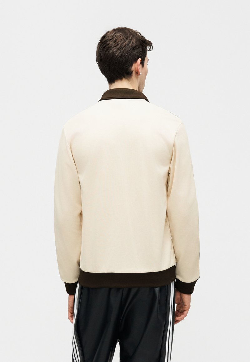 Hombre de espaldas que lleva una chaqueta beige con cuello y puños oscuros, y pantalones negros con rayas verticales blancas en los lados.