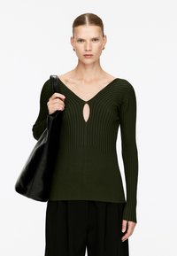 Dunkelgrüner gerippter Pullover mit langen Ärmeln, der einen tiefen V-Ausschnitt und eine Schlüsselloch-Aussparung hat. Kombiniert mit einer schwarzen Schultertasche.