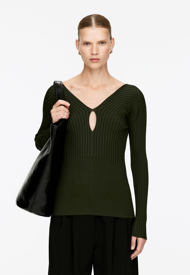Dunkelgrüner gerippter Pullover mit langen Ärmeln, der einen tiefen V-Ausschnitt und eine Schlüsselloch-Aussparung hat. Kombiniert mit einer schwarzen Schultertasche.