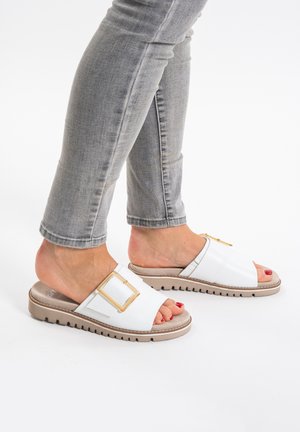 Weiße Rutschen-Sandalen mit einer quadratischen goldenen Schnalle, strukturiertem Obermaterial und einer dicken Sohle. Getragen mit hellgrauen eng anliegenden Jeans.