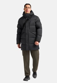 Zwarte pufferjas met capuchon, voorzien van een ritssluiting en schouderriem, gecombineerd met olijfgroene cargobroeken en zwarte schoenen.