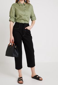 Koszula lniana w kolorze oliwkowej zieleni zapinana na guziki, czarne spodnie z wysokim stanem i skróconą długością, czarna torebka z brązowymi uchwytami, czarne sandały z ozdobną klamrą.