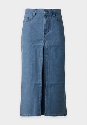 Femme portant un haut blanc à manches longues boutonné et une jupe midi bleue, tenant un grand sac vert olive avec des œillets métalliques ronds, chaussée de talons noirs.