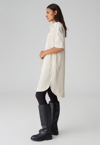 Camicetta abito beige chiaro con maniche corte, chiusura a bottone, fessure laterali, indossata sopra collant neri e stivali alti in pelle nera.
