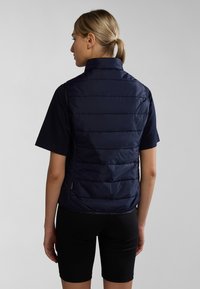 Marinblå väst med vadderad textur och hög krage, med dragkedja och sidofickor, som bärs över en kortärmad skjorta och shorts.