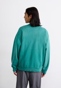 Turkis sweatshirt med en afslappet pasform, ribbede manchetter og kant. Bagfra ses en glat tekstur; iført med grå skræddersyede bukser.