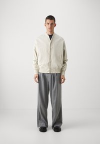 Veste légère couleur crème avec un devant boutonné, des poignets élastiques et deux poches latérales, associée à un pantalon gris large et des chaussures noires.