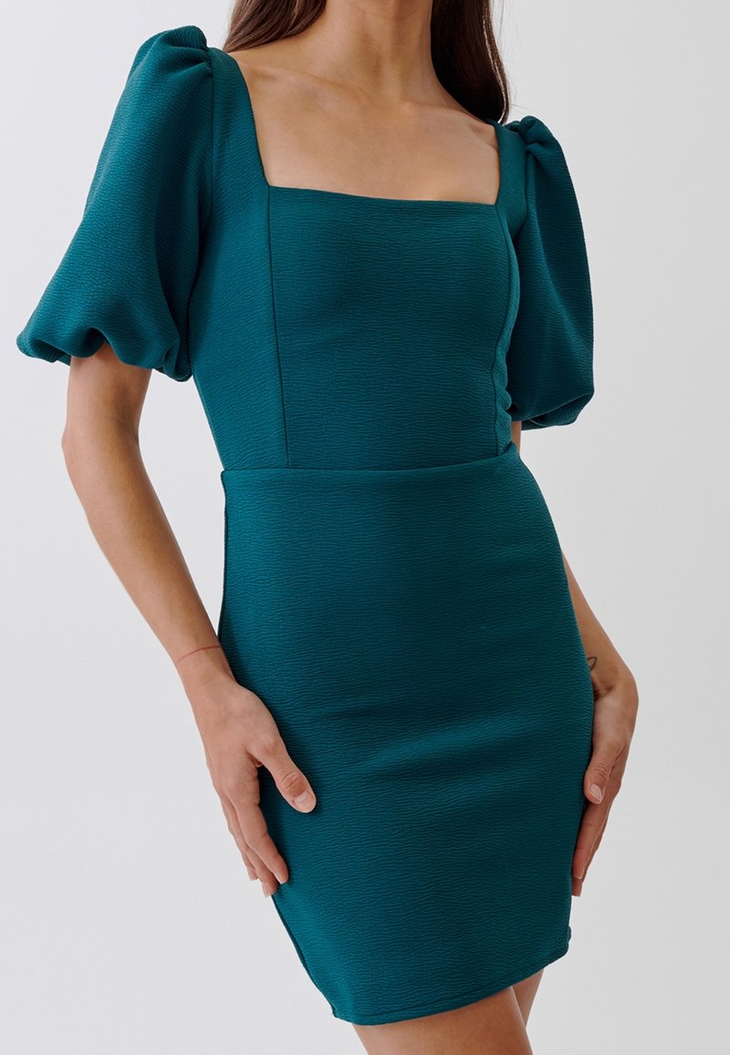 Femme portant une robe ajustée texturée de couleur turquoise avec des manches courtes bouffantes et un décolleté carré, debout les mains posées sur les hanches.