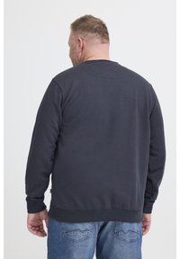 Rövid hajú férfi sötét navy pulóverben és kék farmerban, háttal állva egy sima fehér háttér előtt.