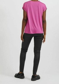 Donna in piedi di spalle, che indossa una maglietta corta rosa brillante, pantaloni neri attillati e scarpe con tacco nero lucido.