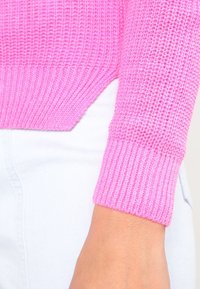 Rosa gestrickter Pullover mit einem strukturierten Muster, geripptem Bündchen und asymmetrischem Saum, kombiniert mit hellblauen Jeans.