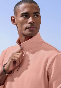 Light peach half-zip pullover made from soft fabric, featuring a structured collar and a silver zipper. The foreground shows a hand adjusting the collar.