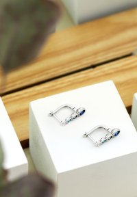 Silverfärgade hängande örhängen med tre runda stenar: två blå och en teal, inramade i en texturerad, bezel-liknande design. Visas på en vit piedestal.