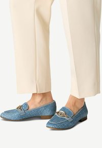 Loafers împletiți de culoare albastră cu tocul flat, având un trim gri și un accent din metal auriu pe partea superioară. Asortați cu pantaloni de culoare crem.