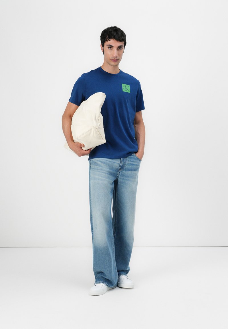 Niebieska bawełniana koszulka z zielonym logo, w połączeniu z jasnoniebieskimi dżinsami, trzymając kremową torbę. Prosty design, casualowy styl.