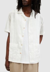 Camisa blanca de manga corta con un patrón texturizado, con botones de madera y dos bolsillos frontales. Llevada sobre una camiseta de color gris claro.