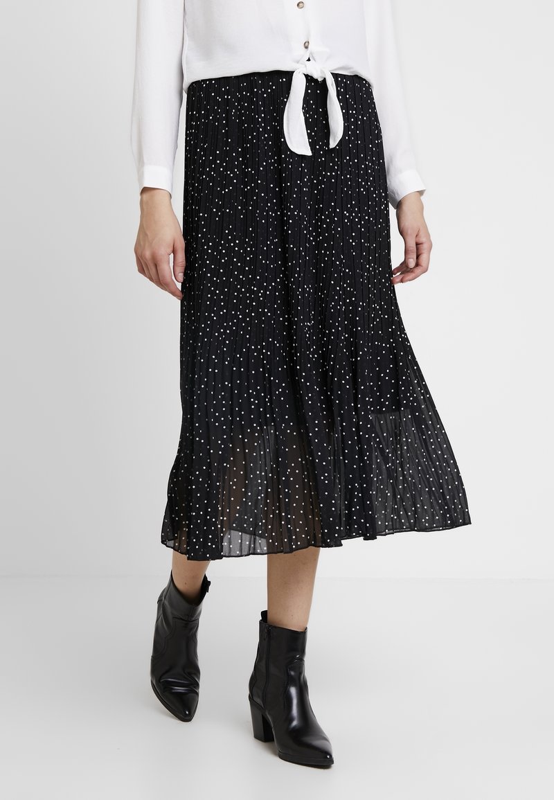 Femme portant une jupe midi plissée noire à pois blancs, un chemisier blanc noué et des bottines à talons noires, debout sur un sol blanc.