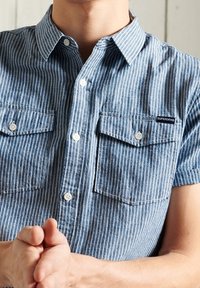 Blue and white striped short-sleeve shirt with button-down collar, two chest pockets, and brand logo on the left pocket. Cotton material.
