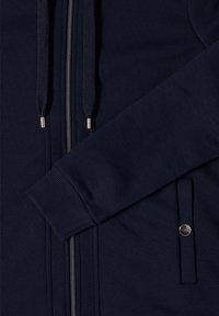 Haut à capuche zippé bleu marine en tissu texturé, avec des poignets côtelés, une fermeture éclair en métal et des cordons de serrage avec des embouts argentés. Comprend une poche latérale.