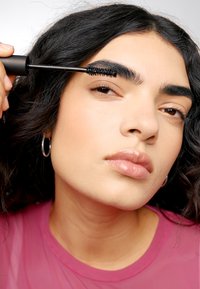 Schwarze Wimperntusche wird auf dicke, dunkle Augenbrauen aufgetragen. Die Person hat lockiges schwarzes Haar und trägt ein pinkfarbenes Oberteil. Der Hintergrund ist sanft verschwommen.