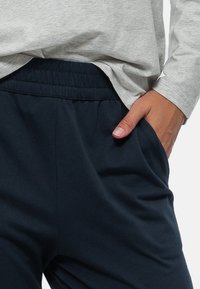 Grå långärmad skjorta över mörkblå, mjuka joggers i tyg med elastisk midja. Hand placerad i joggerfickan.