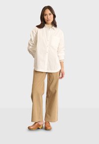 Witte overhemd met knopen en een zak, gecombineerd met tan kleurige wijdvallende corduroy broek en bruine klompen met messing accenten.