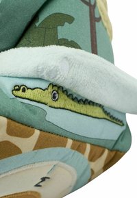 Jucăria din pluș prezintă un design de aligator verde pe un material moale, înconjurat de materiale texturate albastre și bej cu modele de animale și detalii cusute.