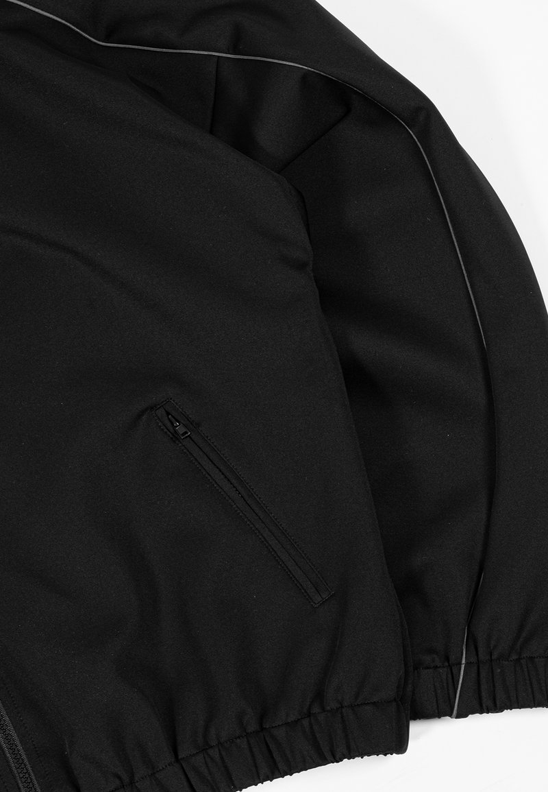 Schwarze Jacke mit seitlichem Reißverschlusstaschen, elastischem Saum und reflektierenden Paspeln entlang der Ärmelkante auf weißem Hintergrund.