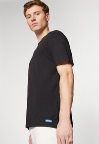 T-shirt nera in cotone con maniche corte, scollatura rotonda, spacchi laterali e logo blu "HUGO" in evidenza sull'orlo. Design minimalista.