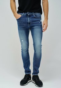 Vaqueros de mezclilla azul con corte ajustado, con desgastes y decoloraciones. Combinados con zapatillas deportivas negras sobre un fondo neutro.