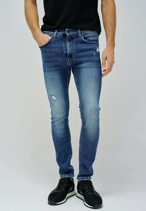 Jean en denim bleu avec une coupe slim, présentant des effets de décoloration et d'usure. Associé à des chaussures de sport noires sur un fond neutre.