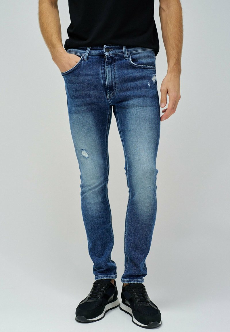 Vaqueros de mezclilla azul con corte ajustado, con desgastes y decoloraciones. Combinados con zapatillas deportivas negras sobre un fondo neutro.