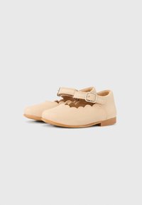 Ζευγάρι μπεζ παπουτσιών Mary Jane για νήπια με κυματιστά άκρα, ρυθμιζόμενες λουρίδες με αγκράφα και επίπεδες καφέ σόλες σε ουδέτερο φόντο.