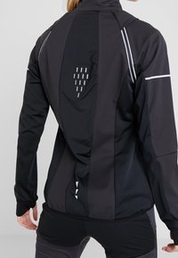 Personne portant une veste de sport noire et gris foncé avec des bandes réfléchissantes et des détails de ventilation au dos, vue de dos.