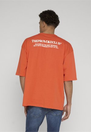 Mann mit lockigem Haar, von hinten, trägt ein orangefarbenes T-Shirt mit weißem Text: „THE PROVOKE CLUB: Erfolg ist die beste Rache, mach es mit Leidenschaft."