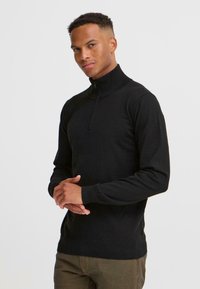 Un jeune homme en sweat à capuche noir se tient confiant, les mains jointes, devant un fond blanc uni.