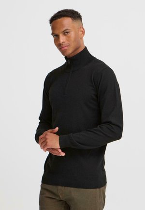 Ein junger Mann in einem schwarzen Reißverschluss-Pullover steht selbstbewusst mit gefalteten Händen vor einem schlichten weißen Hintergrund.