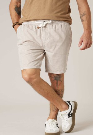 Homme portant un short à cordon beige et blanc à rayures, une chemise marron, des baskets blanches avec des détails noirs, et des tatouages visibles sur les bras et les jambes.